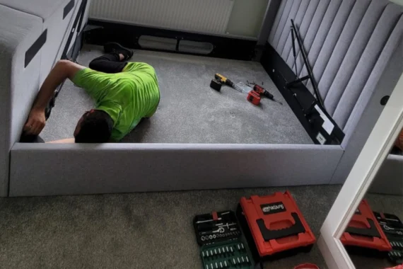 A Mepro Removals expert providing a professional bed assembly service in a Manchester home, fitting a side rail to a grey upholstered bed.