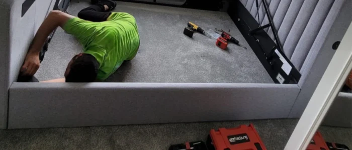 fgsdfgsdfgsdfgsdfgsdf A Mepro Removals expert providing a professional bed assembly service in a Manchester home, fitting a side rail to a grey upholstered bed.