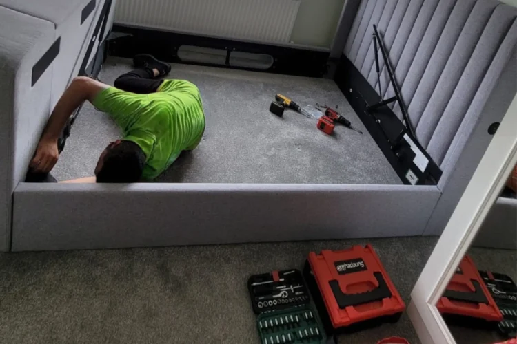 A Mepro Removals expert providing a professional bed assembly service in a Manchester home, fitting a side rail to a grey upholstered bed.