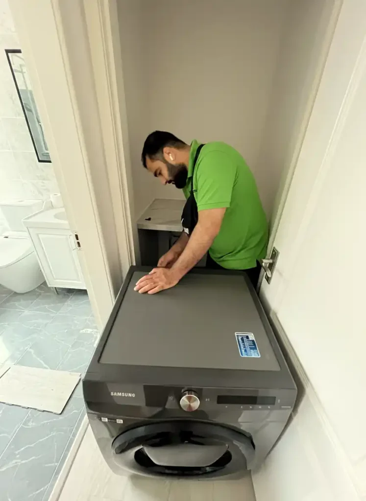 A Mepro Removals expert carefully preparing a Samsung washing machine for moving in a tight Manchester utility space, disconnecting hoses.